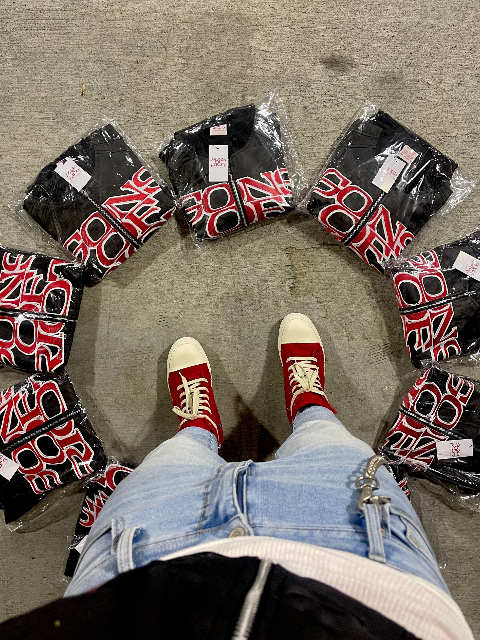 Person wearing red sneakers with white soles standing on a concrete floor surrounded by black jackets with red patterns.